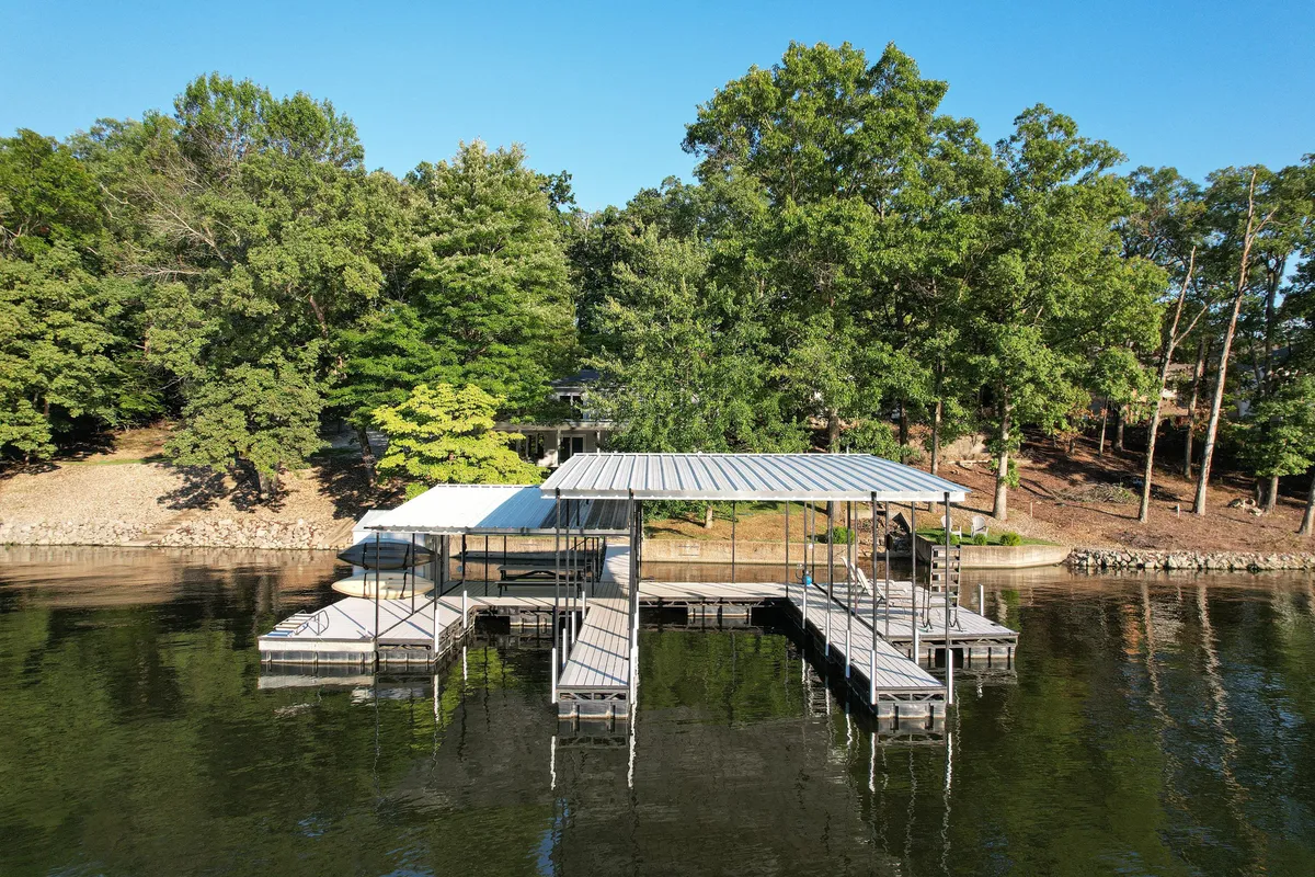 A wide shot of the private dock extending over the calm lake, surrounded by trees with the house visible in the background. The dock is equipped for boating, fishing, swimming, and gathering, making it the centerpiece for water-based fun.