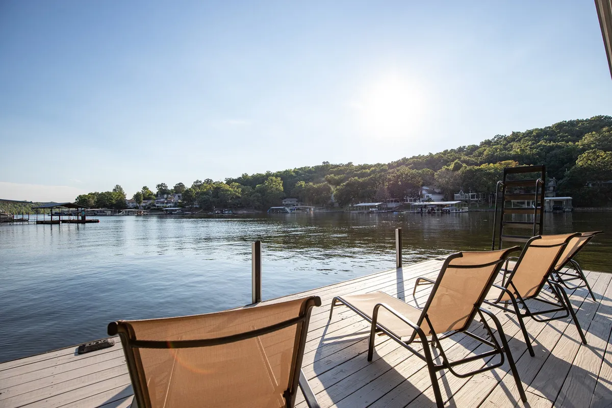 The image showcases the lakefront exterior of the home, with inviting lounge chairs arranged to overlook the serene water and distant rolling hills. The scene is bathed in gentle morning light, hinting at a peaceful sunrise. There’s ample space for relaxing, sunbathing, or gathering with friends and family while enjoying unobstructed views of the lake.
