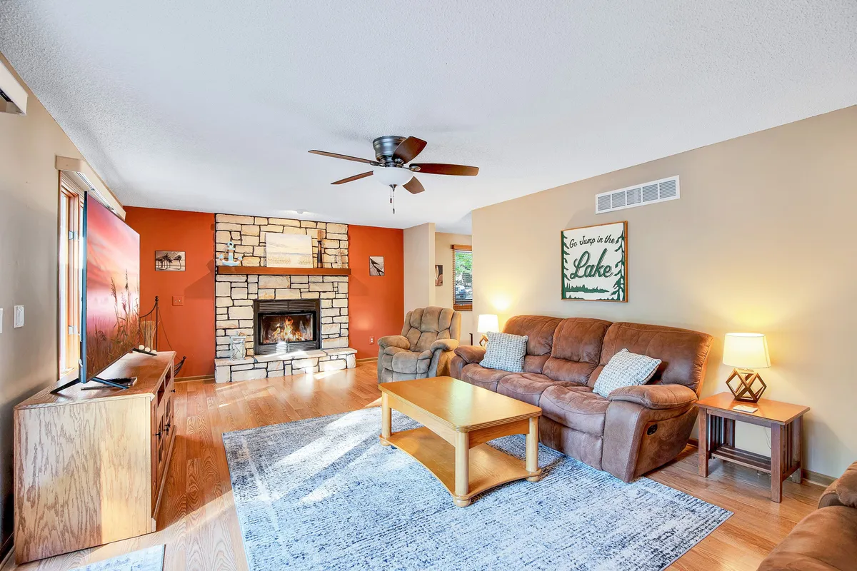 This image captures a spacious living room featuring a stone fireplace, comfortable brown sofas arranged for group seating, a wooden coffee table, and lake-themed wall art. Natural light streams in, creating a warm and inviting atmosphere perfect for relaxing or socializing.