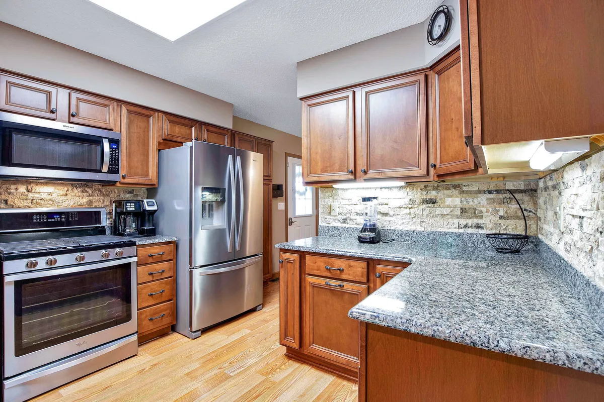 A warm kitchen scene with stainless steel appliances, granite countertops, wooden cabinetry, and a textured stone backsplash. The space radiates a sense of hospitality, ideal for family gatherings and shared meals.