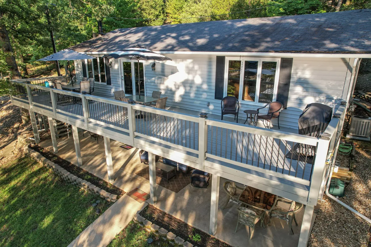 A welcoming exterior shot of the main deck overlooking the lake, featuring comfortable outdoor seating, a large umbrella for shade, and lush greenery surrounding the area. Large windows reflect the natural light, blending indoor and outdoor spaces.