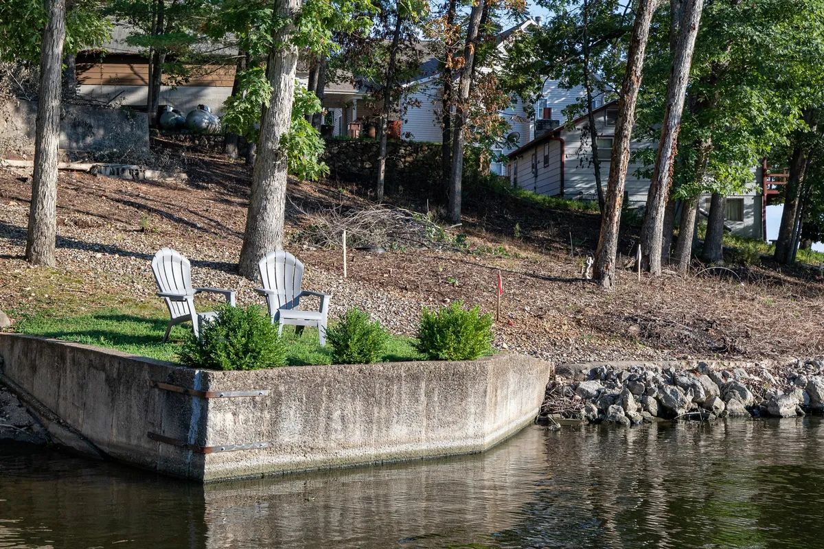 Two classic white Adirondack chairs sit on a concrete dock, surrounded by green shrubbery, facing the calm water. The setting is tranquil, inviting guests to unwind, fish, or watch the sunset in a quiet, natural environment.