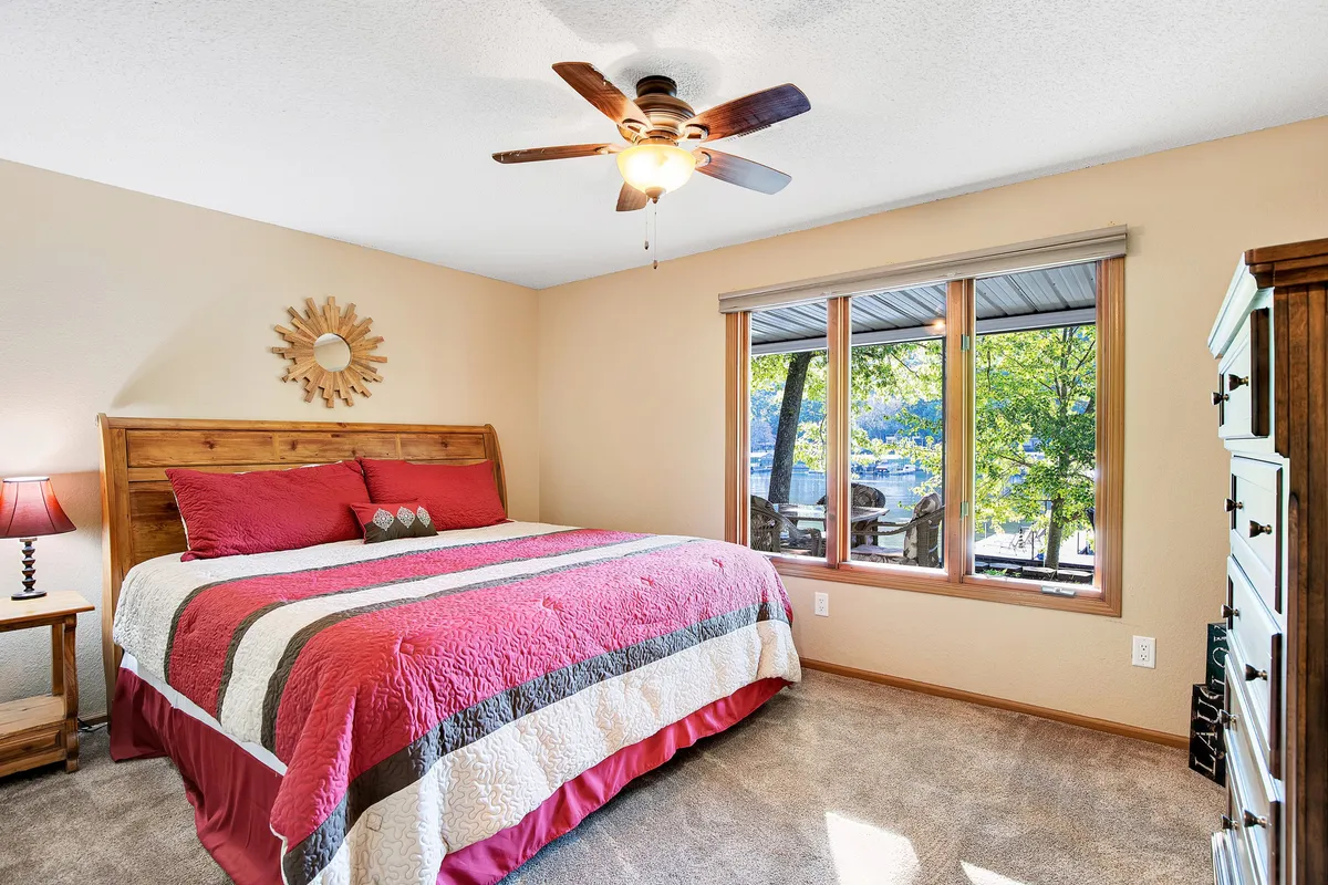 A cozy bedroom featuring a bed with red and white bedding, a large window revealing views of trees and the lake, a nightstand with a lamp, and wooden furniture. Natural light fills the room, creating a tranquil and inviting atmosphere.