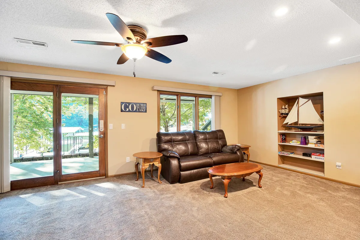 This image showcases a living space with a leather couch, coffee table, bookshelf with decor, and large windows offering views of the outdoors. Ample natural light fills the room, and the arrangement encourages both relaxation and enjoying scenic lake views.