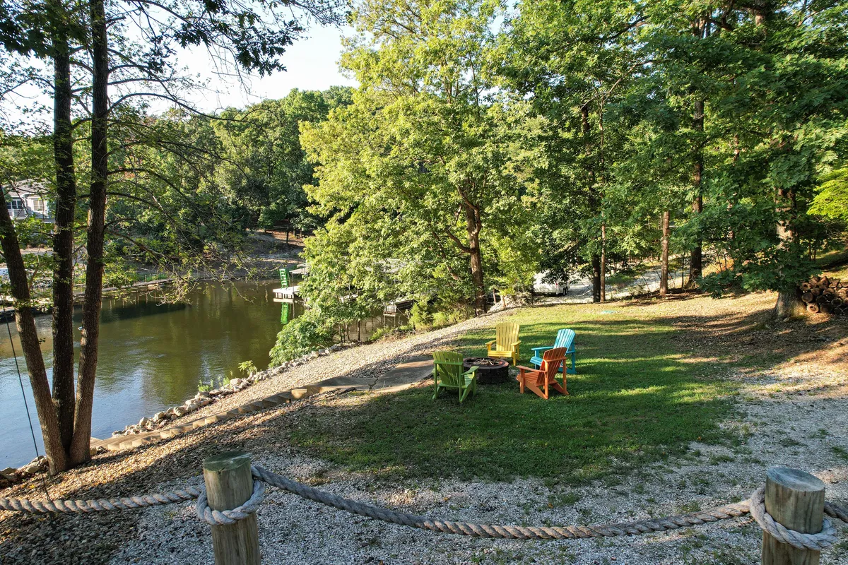 A group of colorful Adirondack chairs arranged around a fire pit, with a scenic water view and trees in the background. The setting is cheerful and relaxed, ideal for gatherings, s'mores, and storytelling by the lake.