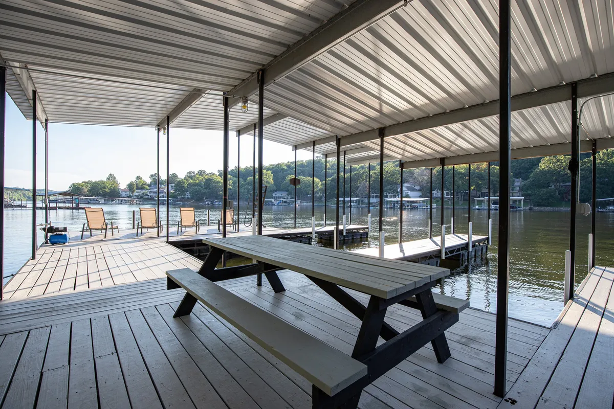 A wide view of the dock area framed by lush trees, featuring a picnic table and lounge chairs overlooking the calm water. The setting showcases space for group gatherings, lakeside meals, fishing, and relaxation by the water, with sunset lighting giving the area a welcoming glow.
